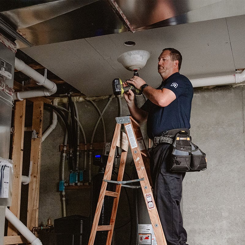 Technician on ladder installing a ceiling fixture in a basement.