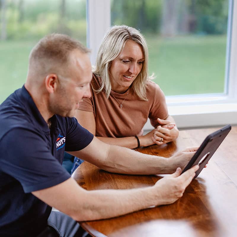 A man and woman sitting at a table looking at a tablet near a window.