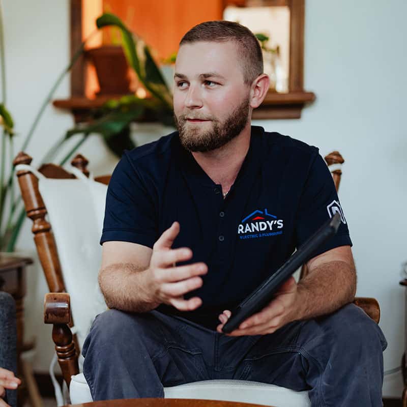 A man in a navy uniform from Summit Electric & Plumbing, holding a tablet, gestures while seated indoors.