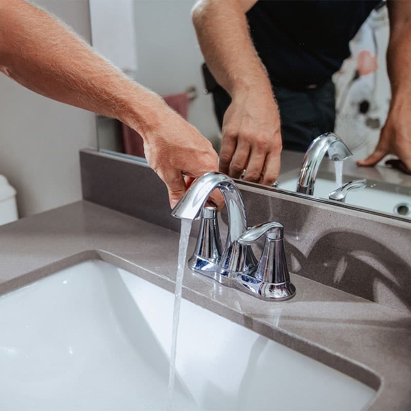 A person turning on a modern chrome bathroom faucet with water flowing into the sink.