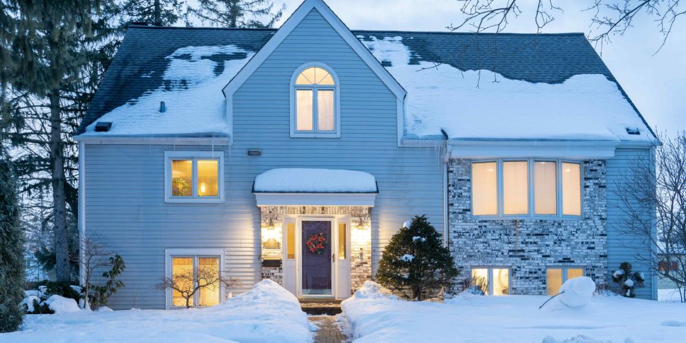 Snow-covered house with lit windows at dusk, surrounded by trees and a path through the snow.