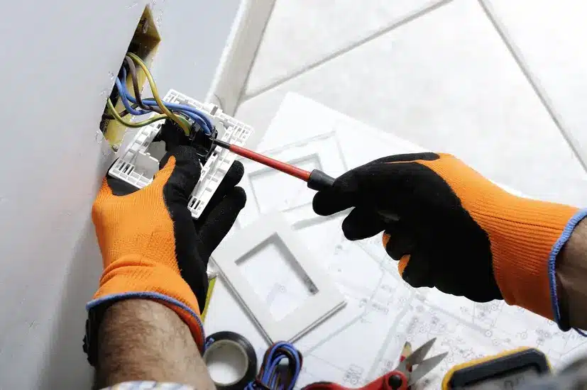 Electrician in orange gloves using a screwdriver to fix a wall socket with exposed wires.