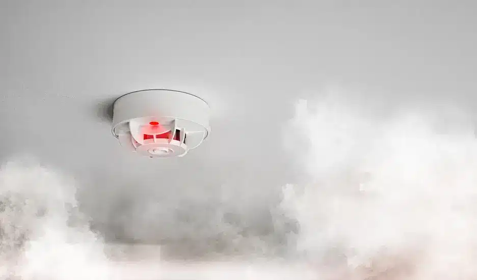 Smoke detector activating amid rising smoke on a ceiling.
