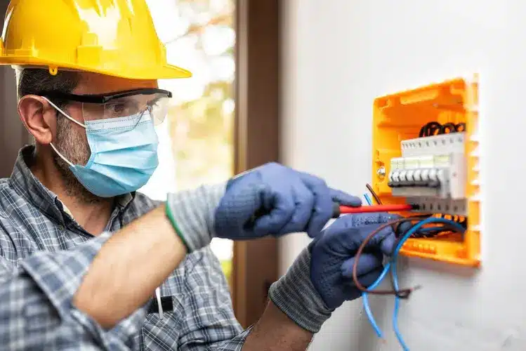 Electrician wearing safety gear repairs electrical panel with tools.