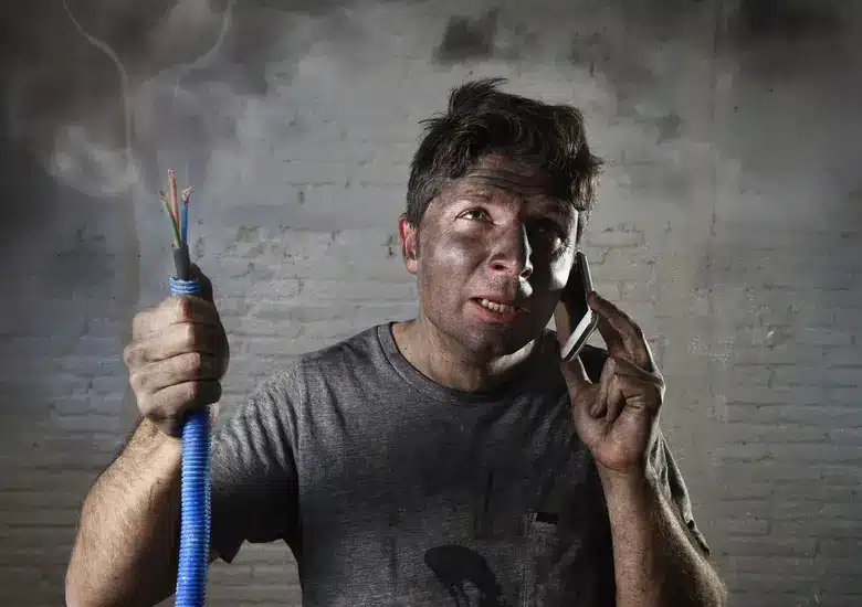 A person with a dirty face holding frayed wires and talking on a phone, looking concerned.