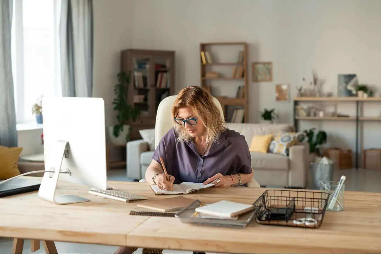 A woman working at a home office desk, writing in a notebook, with a computer and bookshelf in the background.