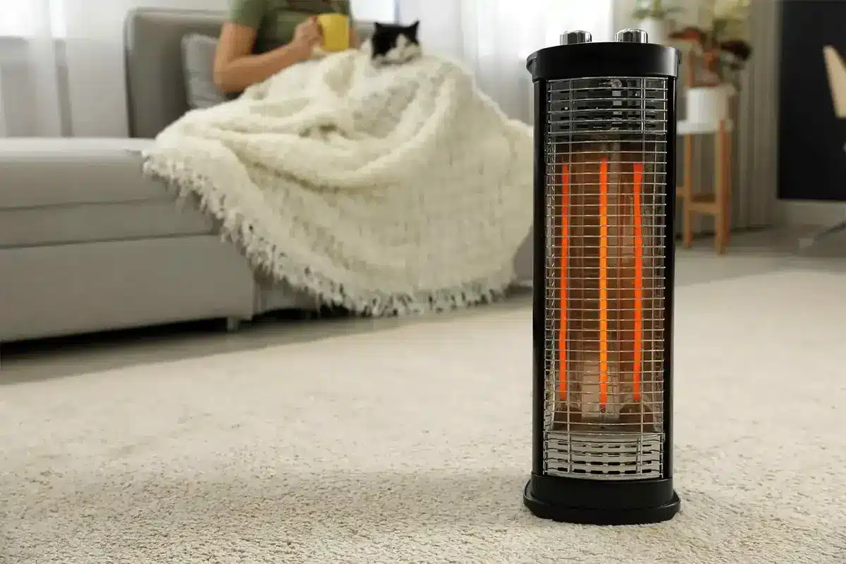 Person on couch with blanket and cat, near electric heater on carpeted floor.