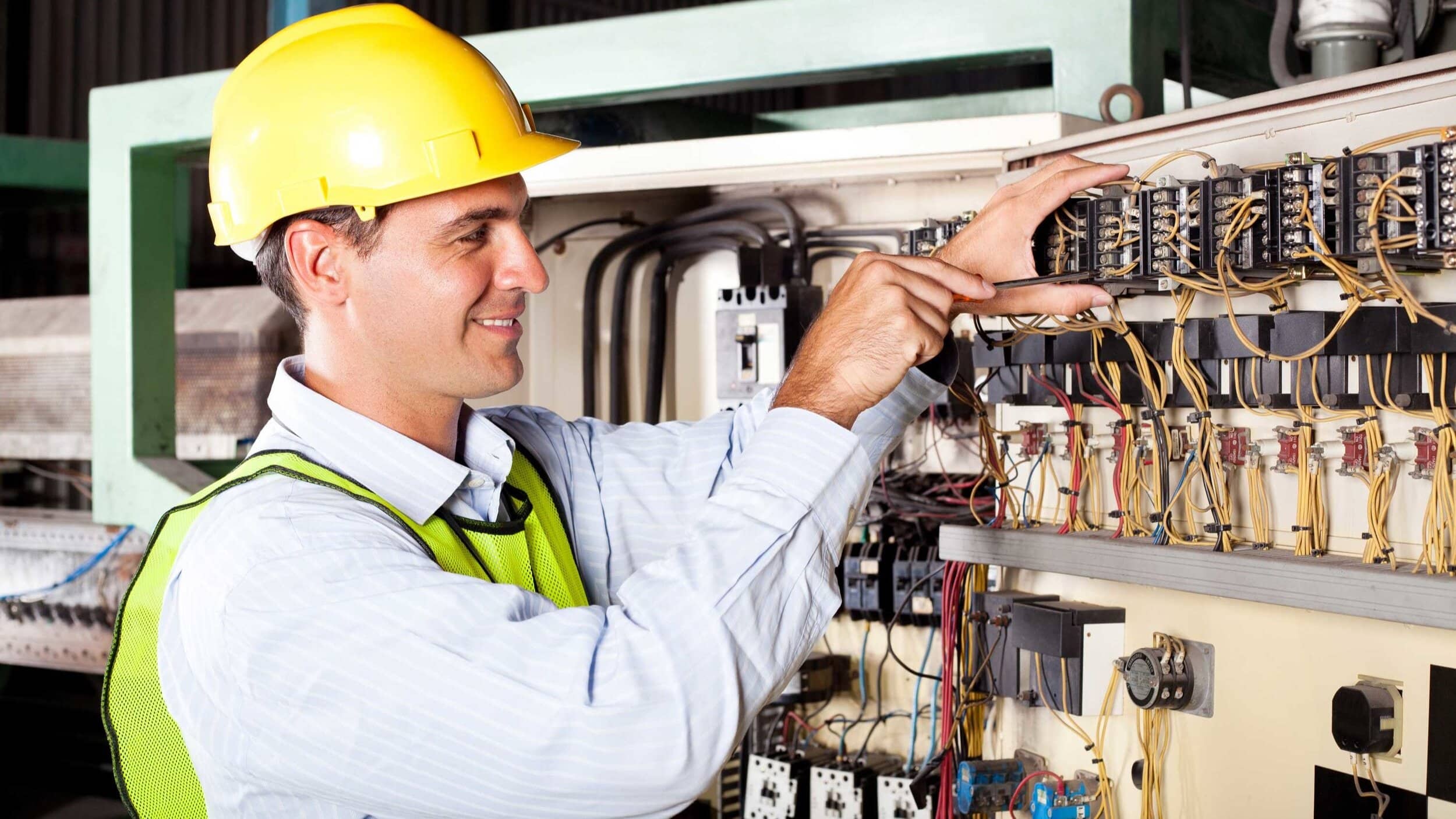 Worker in a hard hat and safety vest, adjusting electrical wiring panel in an industrial setting.