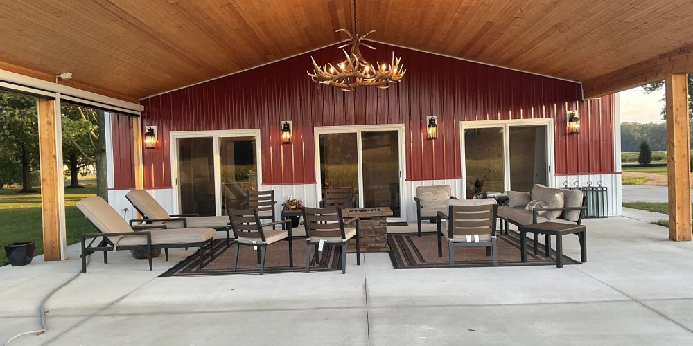 Rustic covered patio at red barn with wood ceiling, antler chandelier, lounge chairs, and fire pit seating