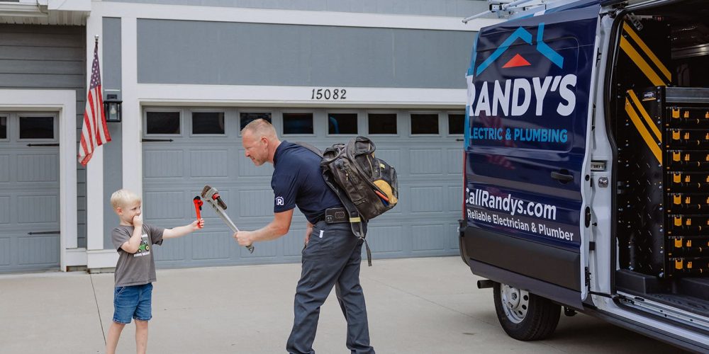 A plumber interacts with a child holding a wrench beside a Summit Electric & Plumbing van in a suburban driveway.