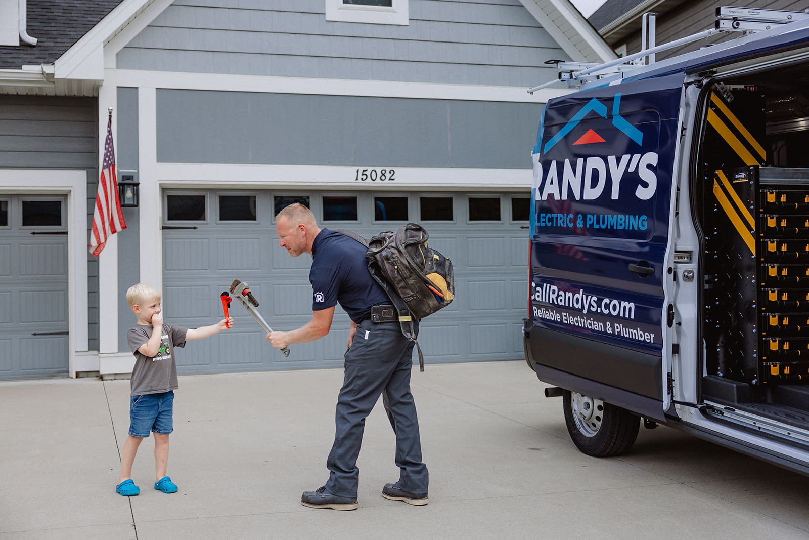 A plumber interacts with a child holding a wrench beside a Summit Electric & Plumbing van in a suburban driveway.